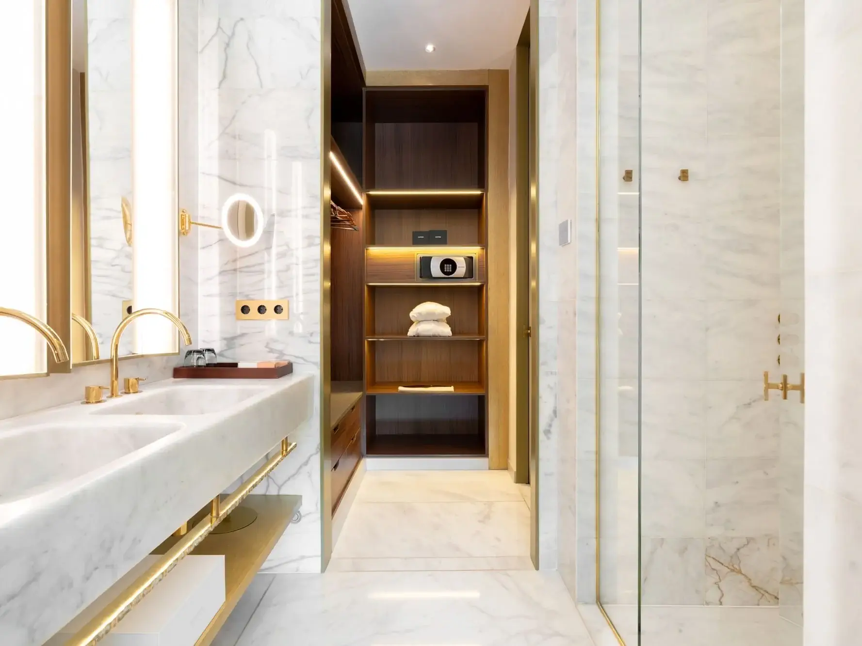 Bany modern de suite familiar amb superfícies de marbre, lavabo doble, accessoris daurats, dutxa de vidre i armari obert de fusta amb prestatges, caixa forta i tovalloles ben plegades: experimenteu el luxe d'Almanac amb cada detall.