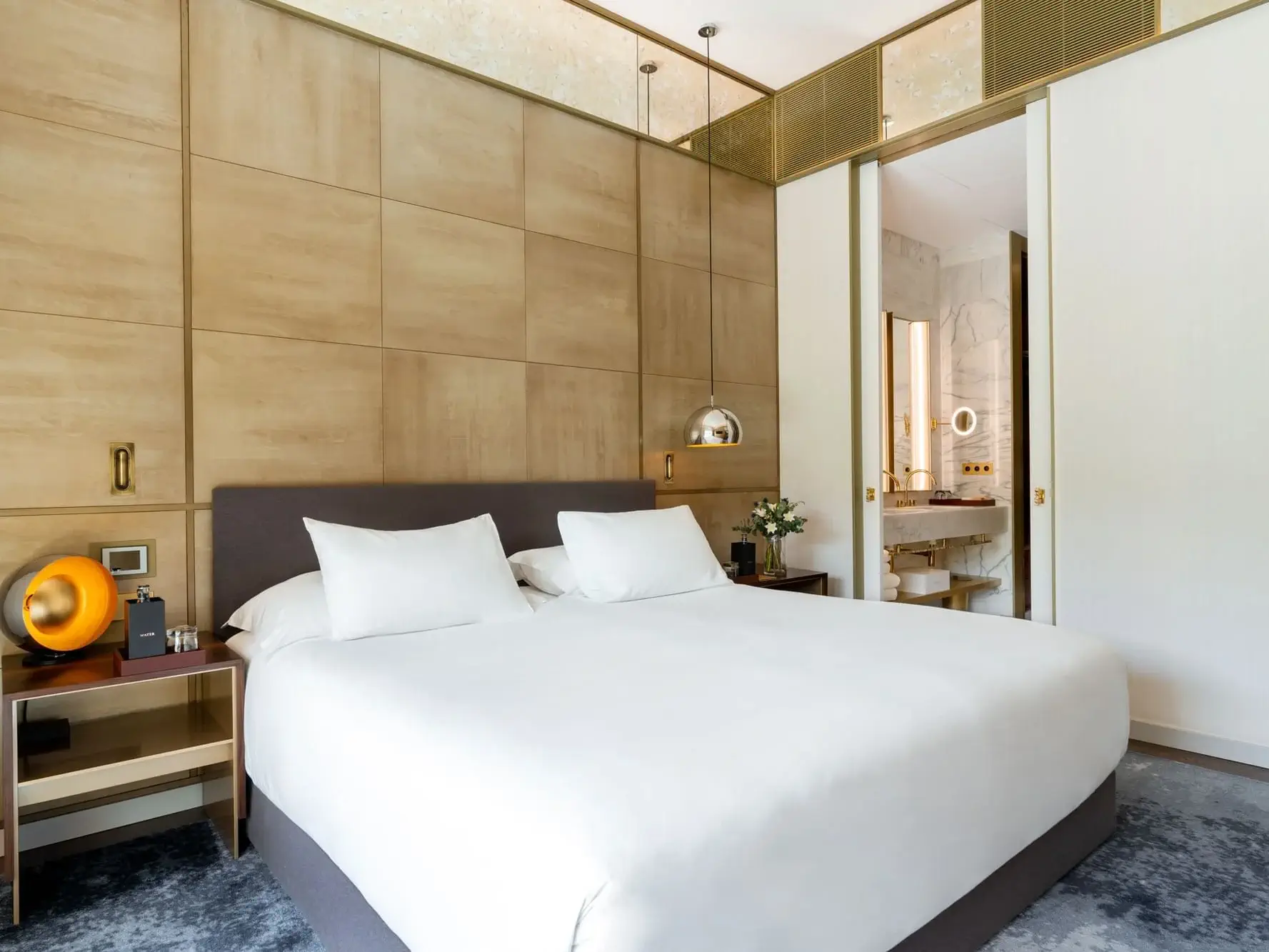 Suite familiar moderna Almanac amb un llit gran, dos coixins blancs, tauletes de nit, panells de fusta càlids i una porta corredissa oberta que revela un bany amb parets de marbre.