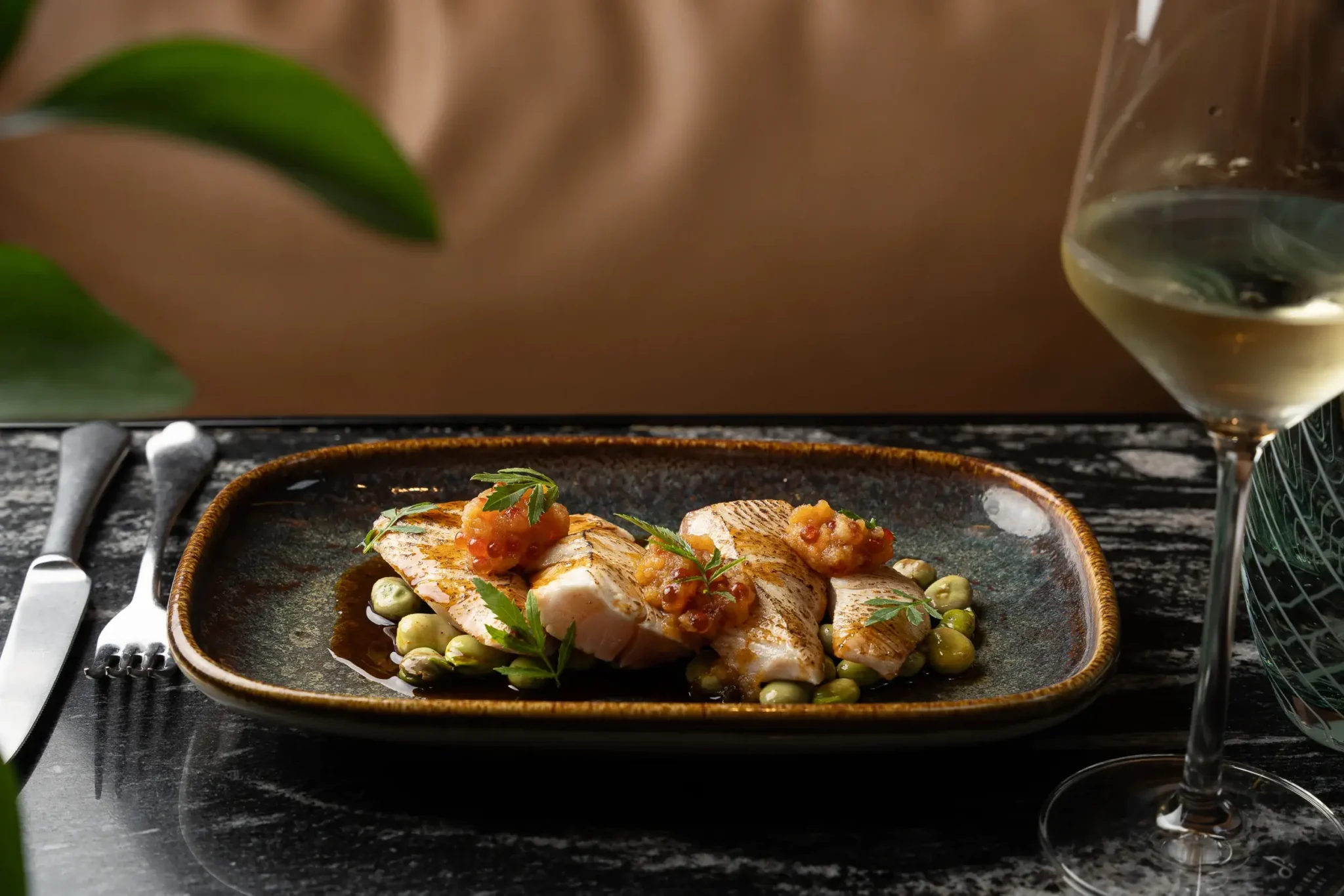 A plated dish of sliced fish with beans and a vegetable garnish sits on a dark table next to a glass of white wine, showcasing an elegant pairing of food and drinks, with cutlery placed neatly beside the plate.
