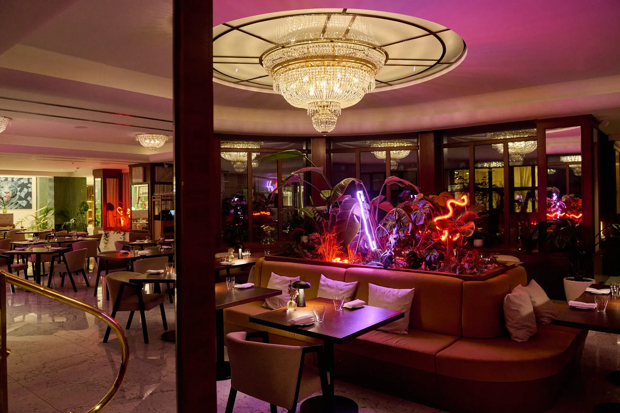 Moderní interiér pražské restaurace s elegantními lustry, prohnutými sedadly, malými jídelními stolky a neonovými světly osvětlujícími rostliny nabízí zážitek z dobrého jídla připomínající Restauraci Alcron.