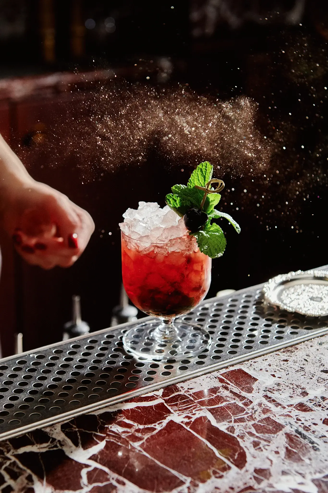 A hand sprinkles powder over a red cocktail with crushed ice, mint, and a cherry garnish, sitting on a bar counter.