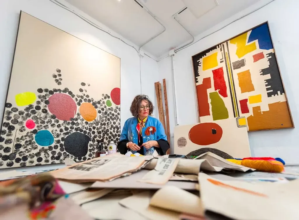 An artist sits in a studio surrounded by abstract paintings and scattered papers, preparing for upcoming art events, with meeting notes, art supplies, and canvases visible around her.