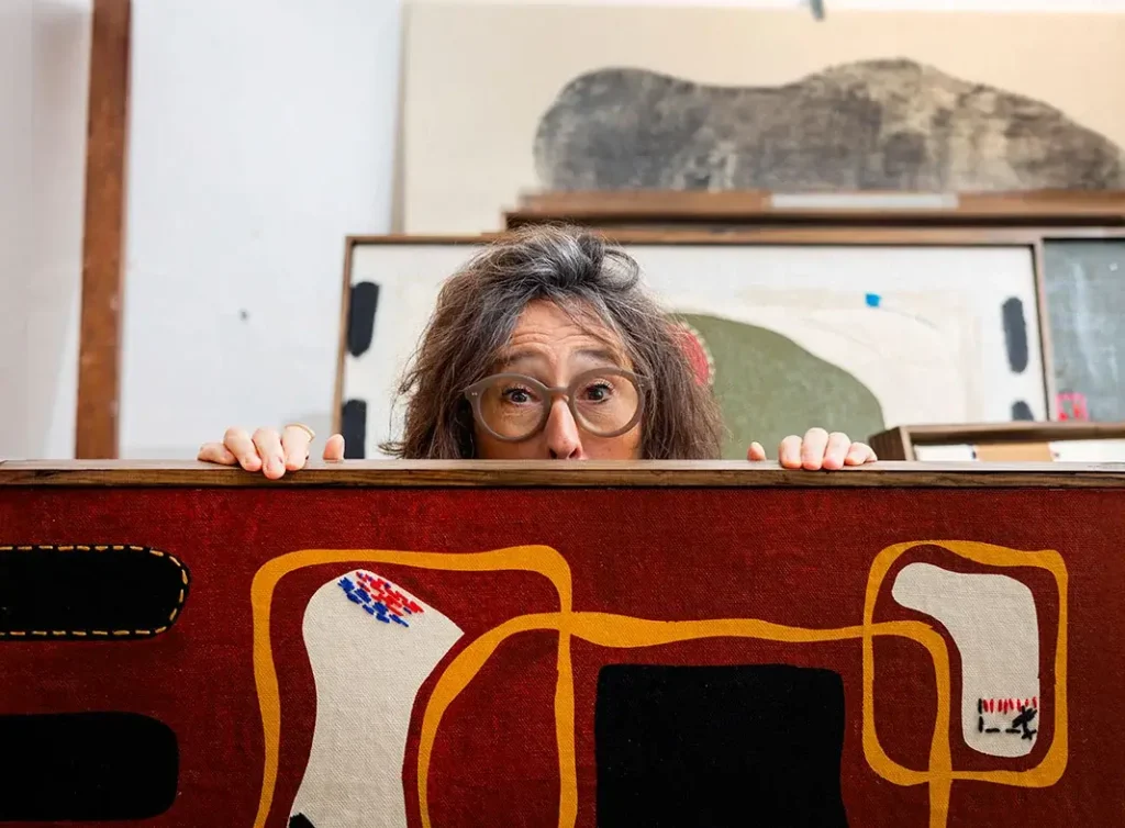A person with glasses peers over a colorful abstract painting in an art studio, preparing for an upcoming art meeting, with several other vibrant artworks displayed in the background.