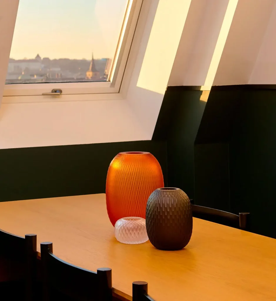 Three textured glass vases—one orange, one black, and one clear—sit on a wooden table near a window with a cityscape view and sunlight streaming in, creating an inviting setting for meetings or intimate events.