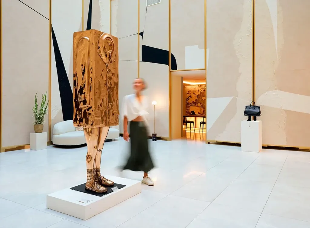 A blurred person walks past a modern golden sculpture of a headless human figure in a minimalist, well-lit gallery space, where abstract wall art sets the scene for exclusive events or an inspiring meeting.