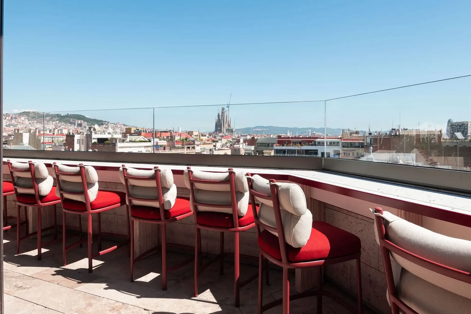 A row of red cushioned bar stools lines the Azimuth Rooftop Bar terrace, overlooking a cityscape with clear skies and distant buildings.
