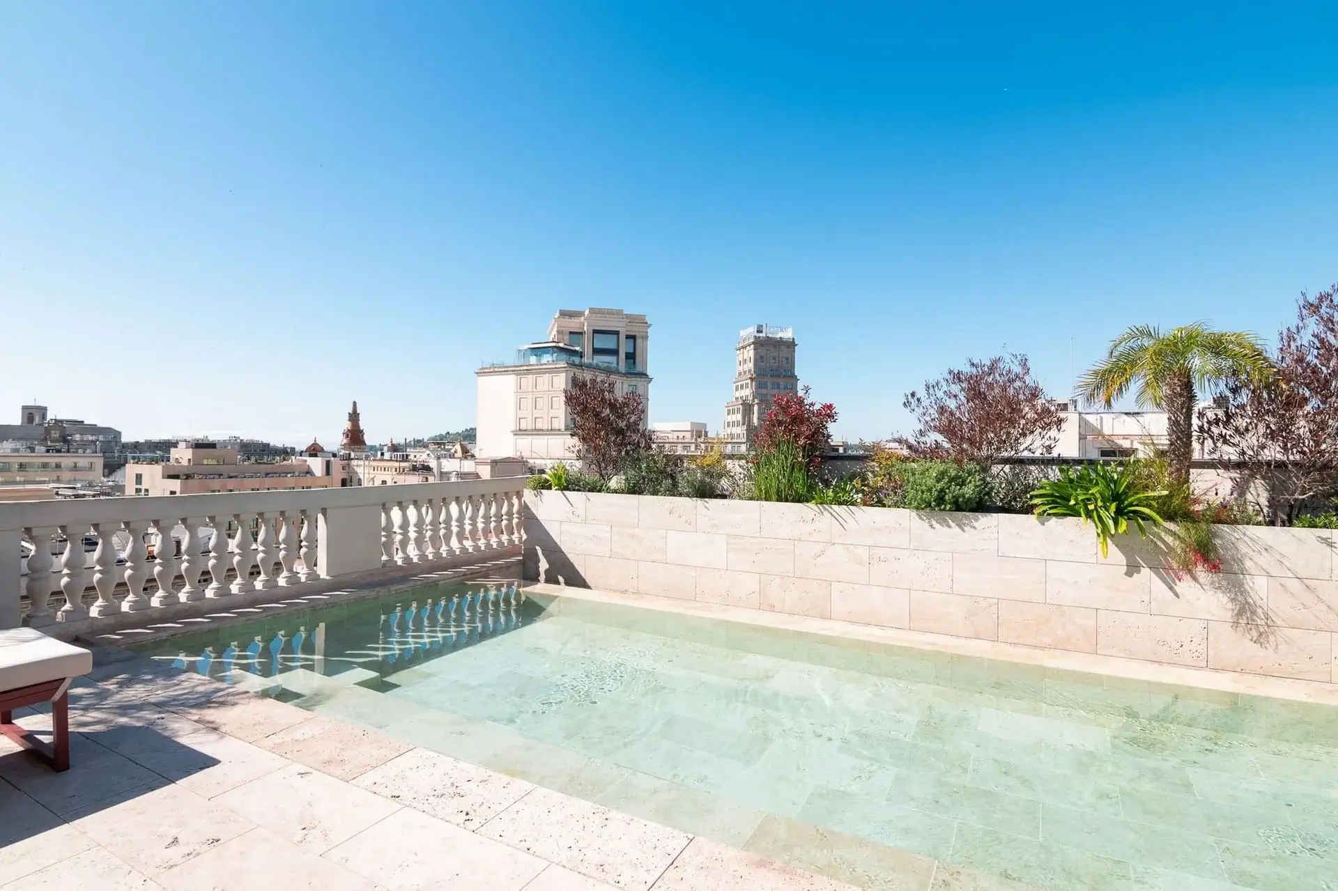 A rooftop terrace at Azimuth Rooftop Bar features a shallow rectangular pool, stone railing, lush plants, and a stunning cityscape in the background under a clear blue sky.
