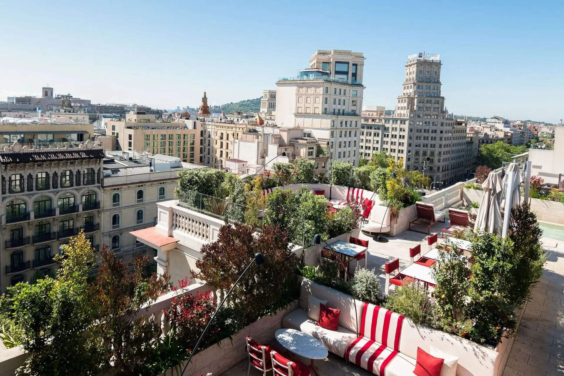 Azimuth Rooftop Bar features red and white striped seating, tables, and lush greenery on a rooftop terrace overlooking a cityscape of historic and modern buildings under a clear blue sky.