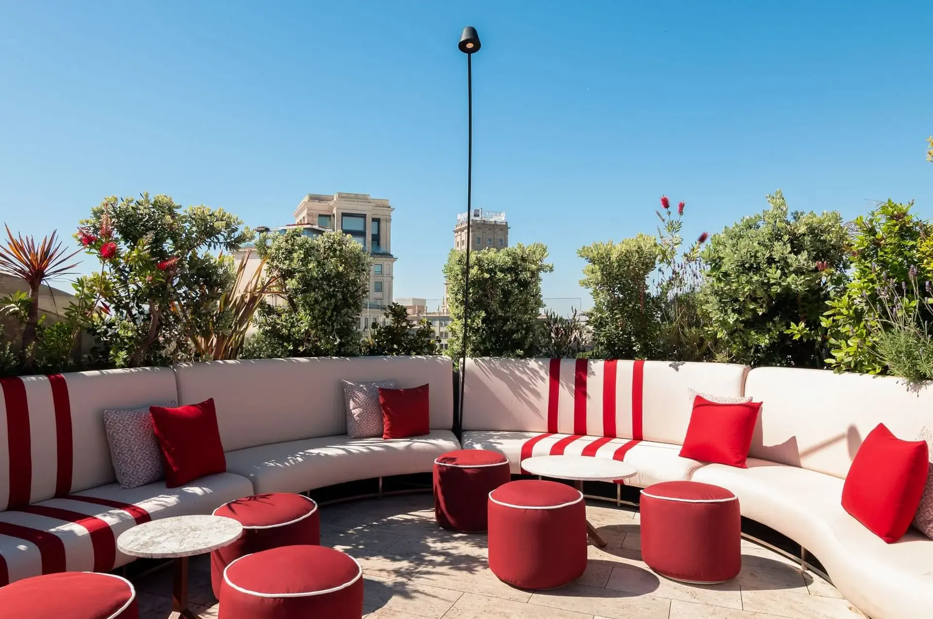 Azimuth Rooftop Bar features curved white seating, red and white cushions, red ottomans, small round tables, lush greenery, and city buildings in the backgroundโall under a clear blue sky.