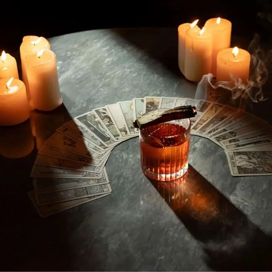 A glass of whiskey with a smoking incense stick rests on a curved spread of tarot cards, surrounded by lit candles on a dark tabletop, inviting a new perspective beneath the mysterious glow—an azimuth of fate revealed.