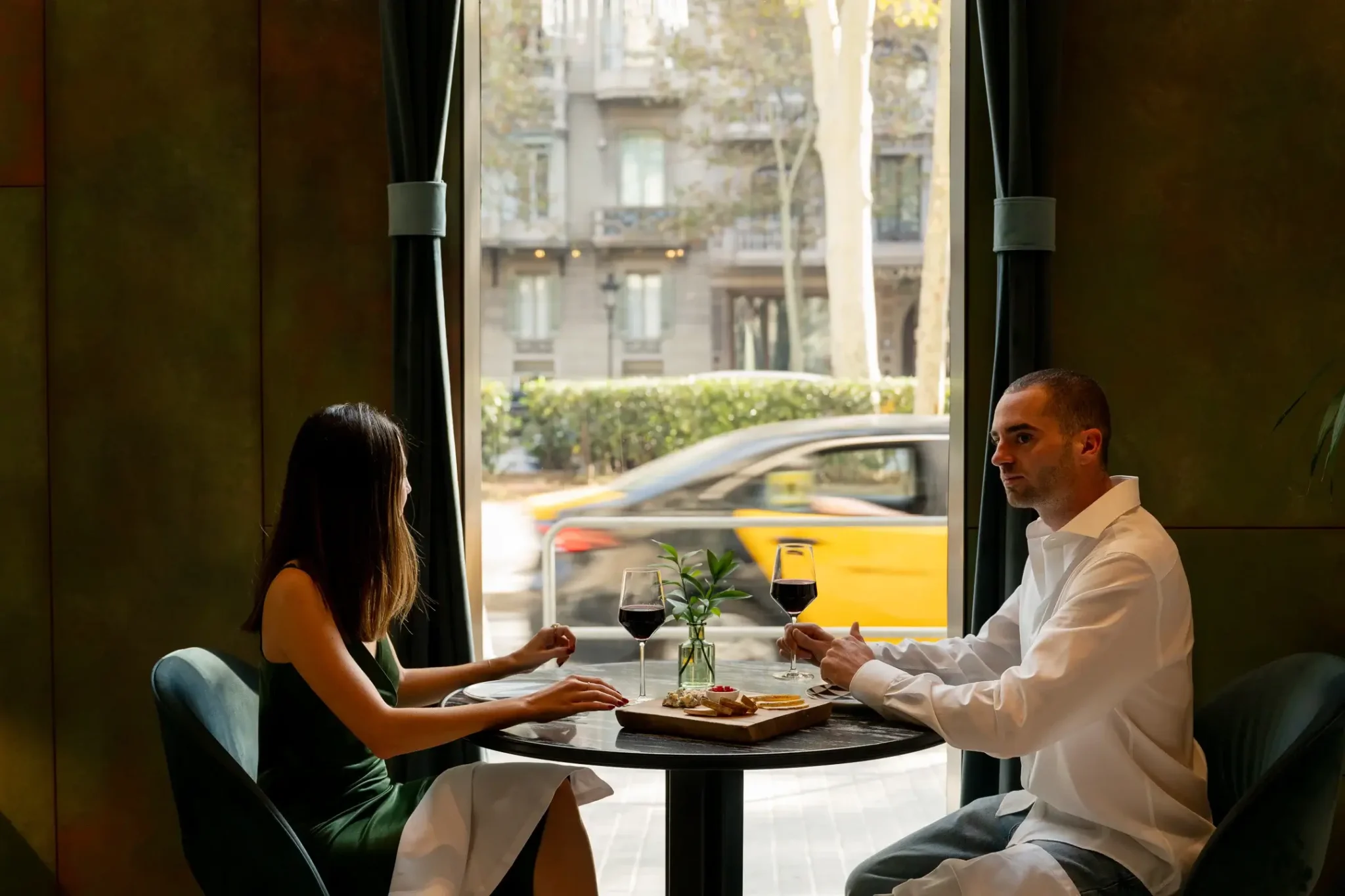 Un hombre y una mujer se sientan en una mesa junto a una ventana, compartiendo vino y aperitivos durante su Escapada de Otoño, mientras un taxi amarillo pasa por fuera, en la calle de la ciudad.