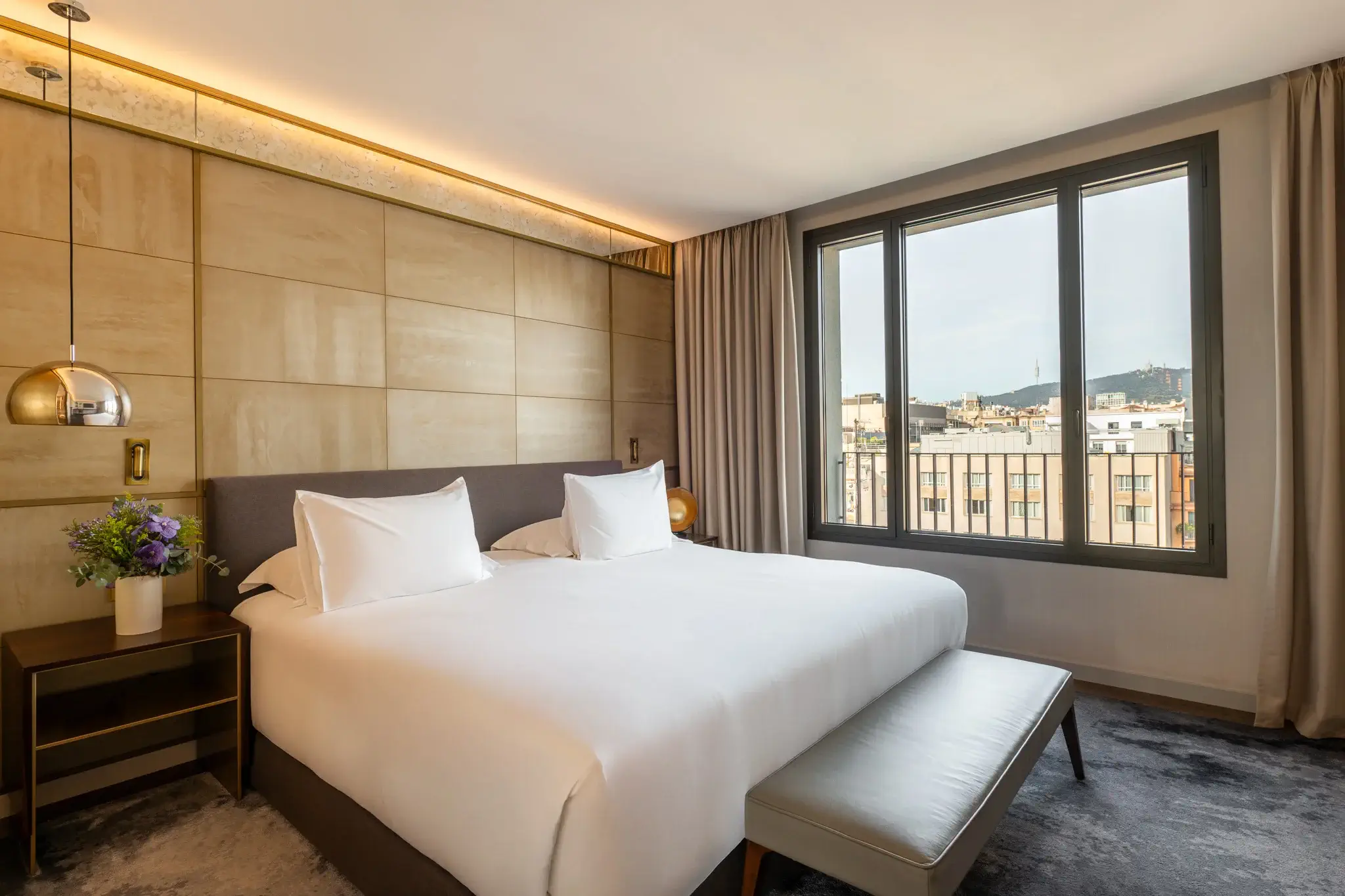 Una Habitació Family moderna amb un llit gran, roba de llit blanca, tauletes de nit amb làmpades, un banc als peus del llit i una finestra amb unes vistes impressionants de la ciutat, ideal per a famílies que busquen comoditat en una habitació d'hotel.