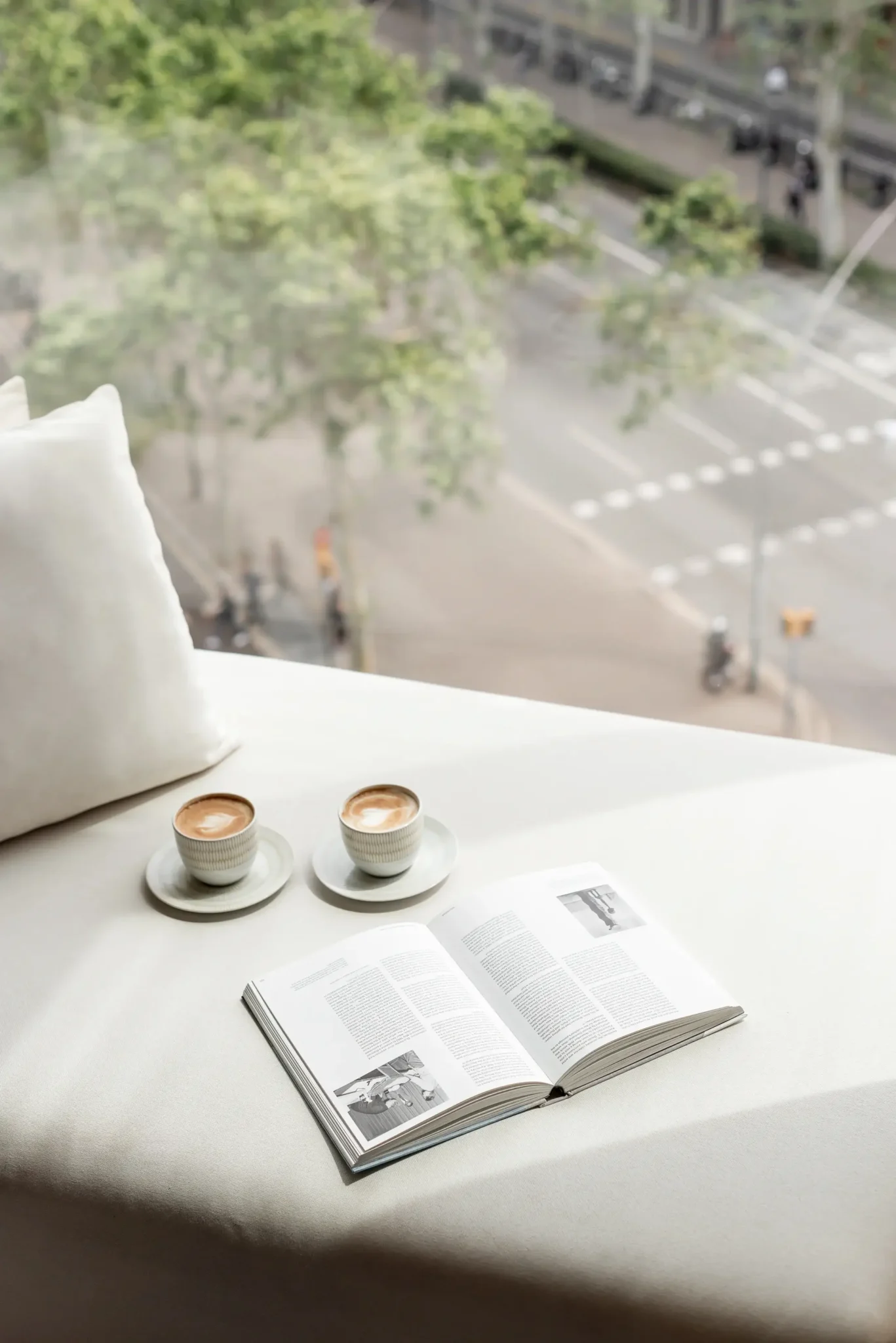 Dos tazas de café y un libro abierto descansan en el alféizar de una ventana blanca, capturando el ambiente perfecto para una Escapada de Otoño, con una calle de la ciudad visible a través de la ventana en el fondo.