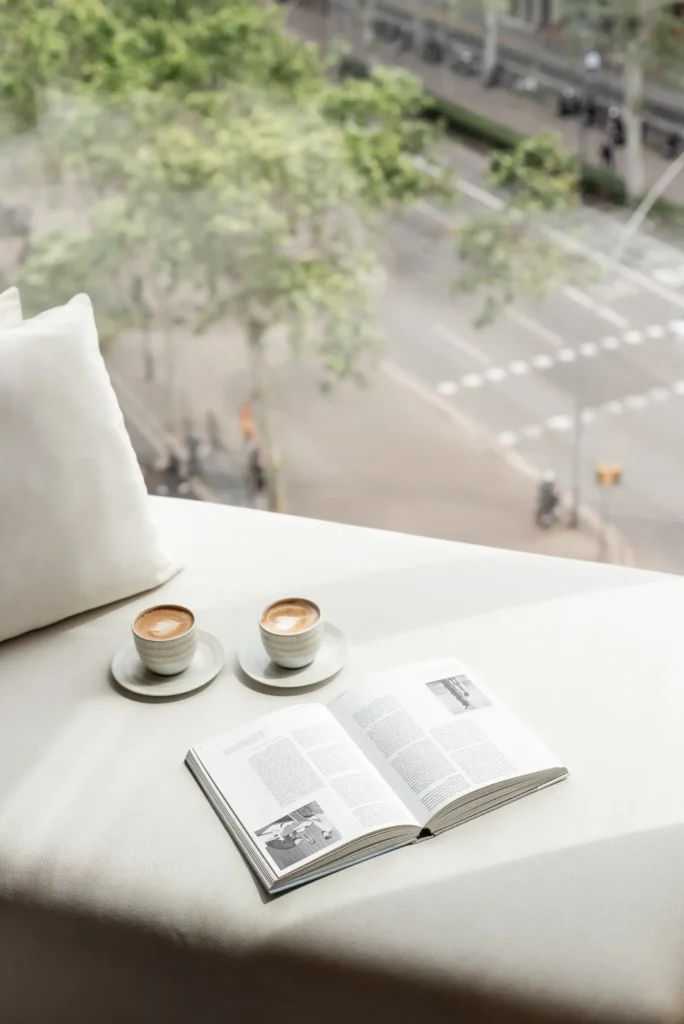 Two cups of coffee and an open book rest on a white surface by a window, offering the perfect autumn escape overlooking a city street lined with trees and traffic.