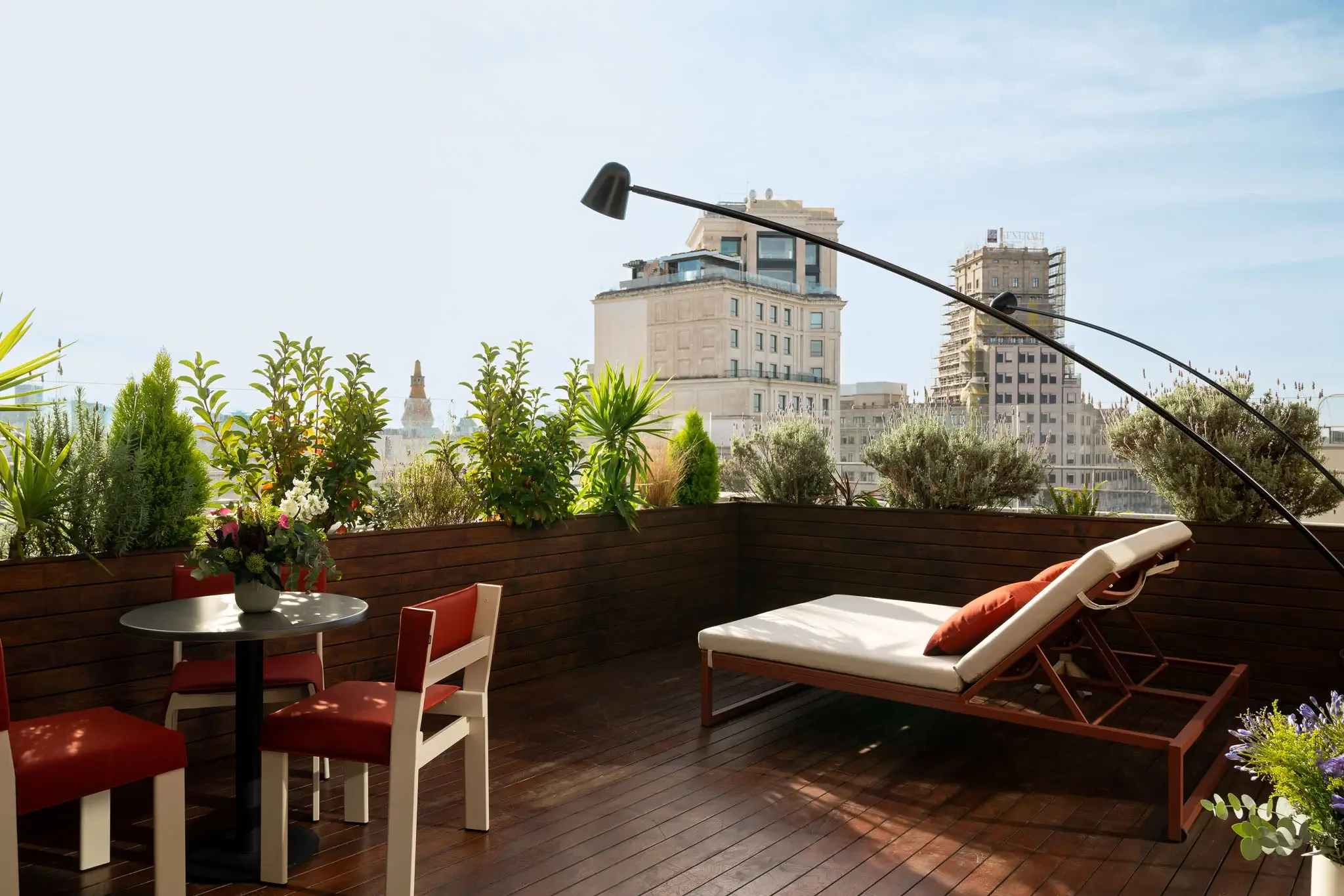 Una terrassa a la teulada d'una suite de luxe compta amb terres de fusta, una butaca, una petita taula amb cadires, plantes en test i edificis de la ciutat al fons sota un cel clar.