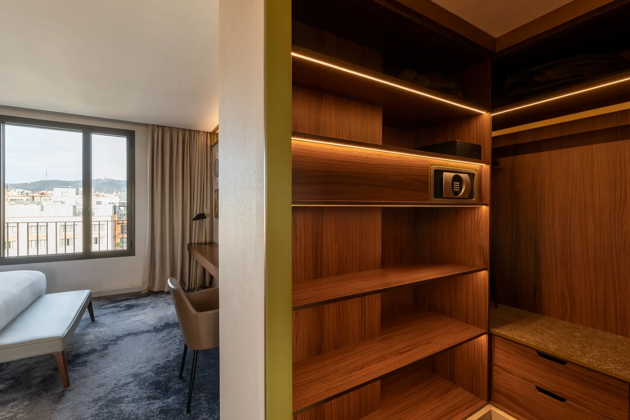 Una habitació d'hotel de luxe tipus suite té un llit i un escriptori a l'esquerra, amb un armari de fusta obert amb prestatges i una caixa forta a la dreta.