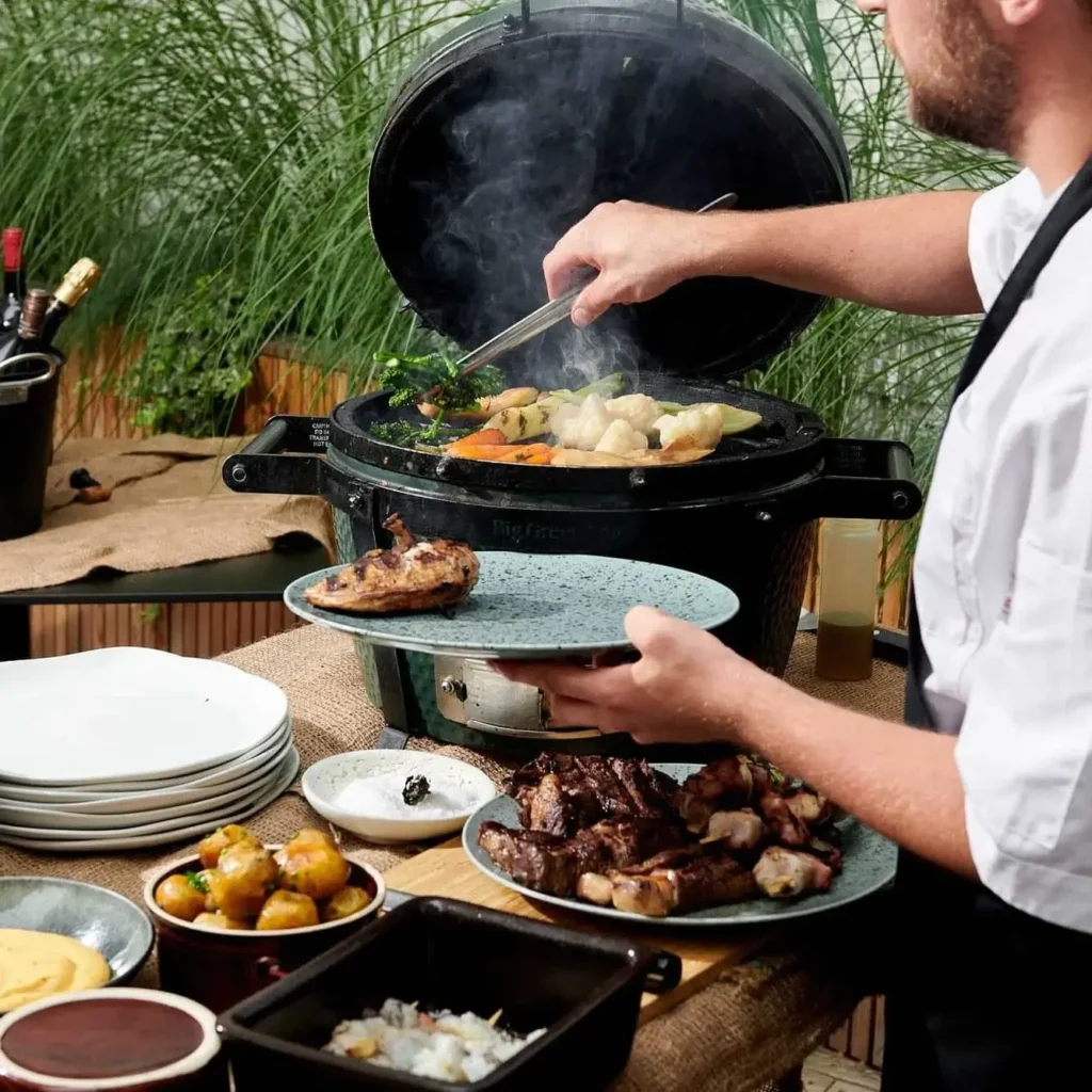 Eine Person grillt Gemüse und Fleisch auf einem Holzkohlegrill für ein BBQ-Mittagessen, wobei verschiedene gekochte Gerichte und Teller auf einem Tisch in der Nähe angeordnet sind.