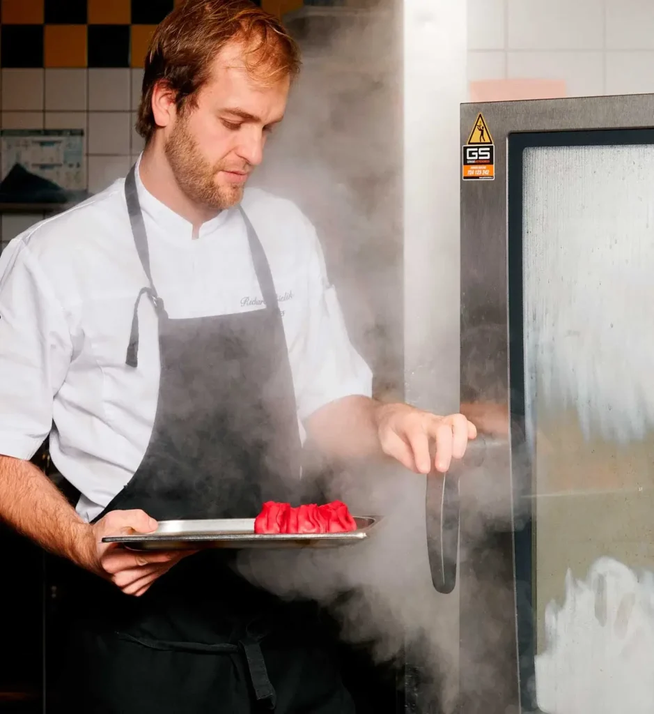 A chef opens a steaming oven while holding a tray with red food items in a commercial kitchen.