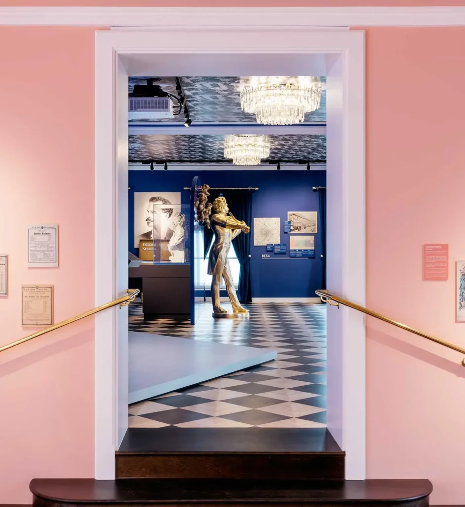 View through a doorway into a gallery with checkered floors, blue walls, framed art, and a gold sculpture at the center; chandeliers hang from the ceiling.