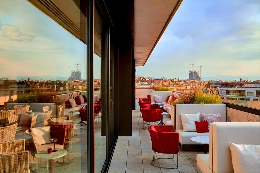 Moderne Dachterrasse mit Korbsesseln, roten Kissen, weißen Sofas, Glaswänden und Blick auf die Skyline der Stadt bei Sonnenuntergang.