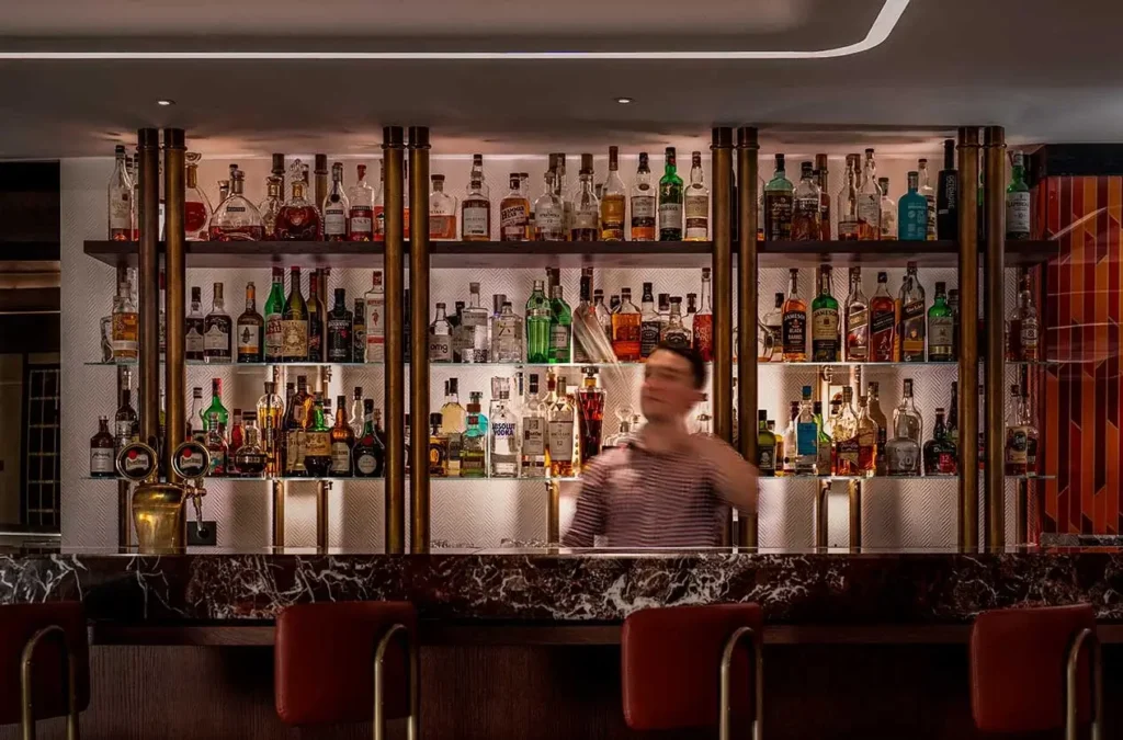Ein Barkeeper steht hinter einer Marmorbar mit roten Stühlen und vor Regalen mit verschiedenen Schnapsflaschen.