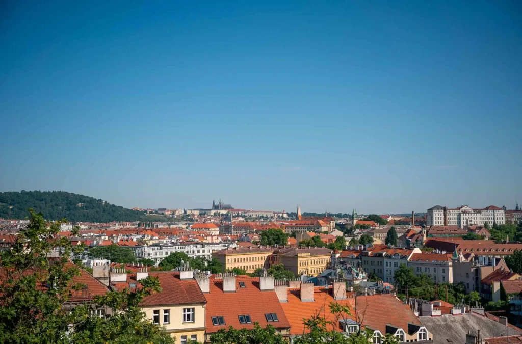 Stadtlandschaft mit roten Dächern, Bäumen und entfernten Hügeln unter einem klaren blauen Himmel an einem sonnigen Tag.