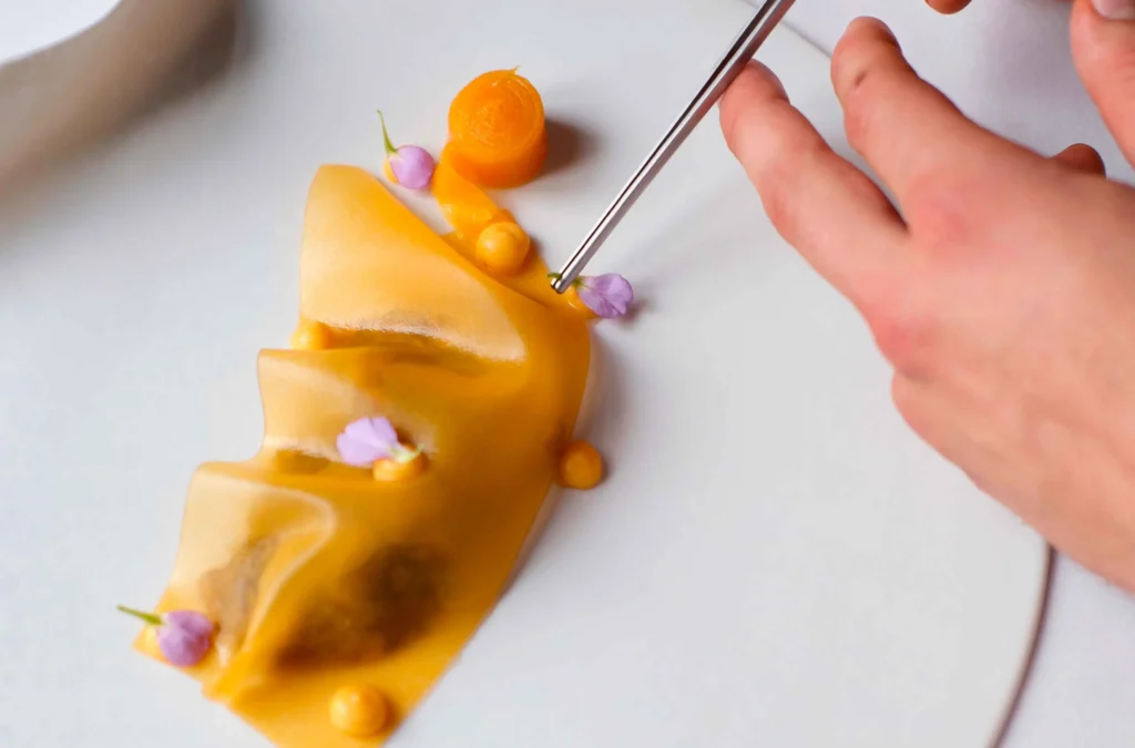 Eine Hand setzt mit einer Pinzette eine violette essbare Blume auf ein Gourmetgericht mit zarter gelber Soße und kleinen Gemüsebeilagen.