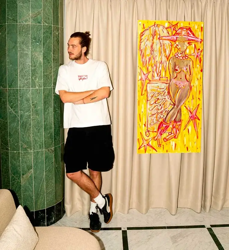 A man in a white t-shirt and black shorts stands by a green marble pillar, near a colorful painting on a curtain backdrop.