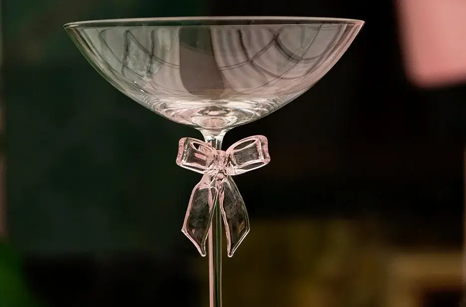 A clear glass cocktail coupe with a decorative glass bow tied around the stem, set against a blurred background.