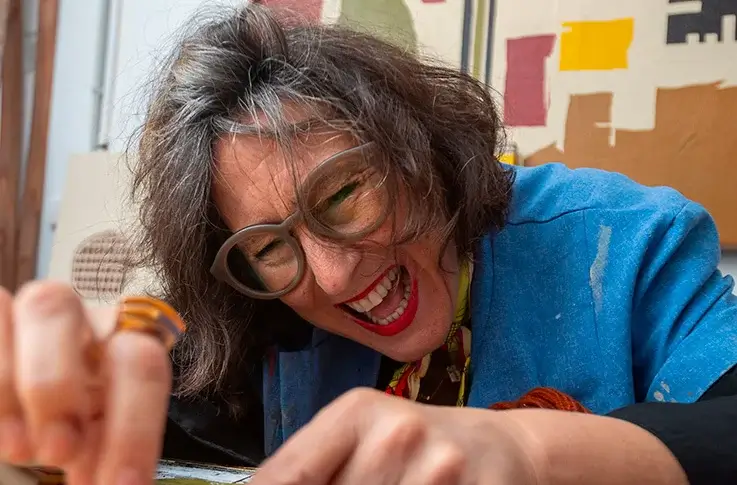 Person with gray-streaked hair wearing glasses and a blue smock is smiling and working closely on an art project in a studio setting.