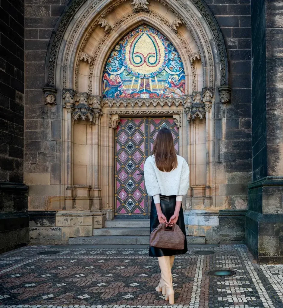A woman with long brown hair stands facing an ornate church doorway with colorful stained glass and detailed stonework, holding a brown bag behind her back.