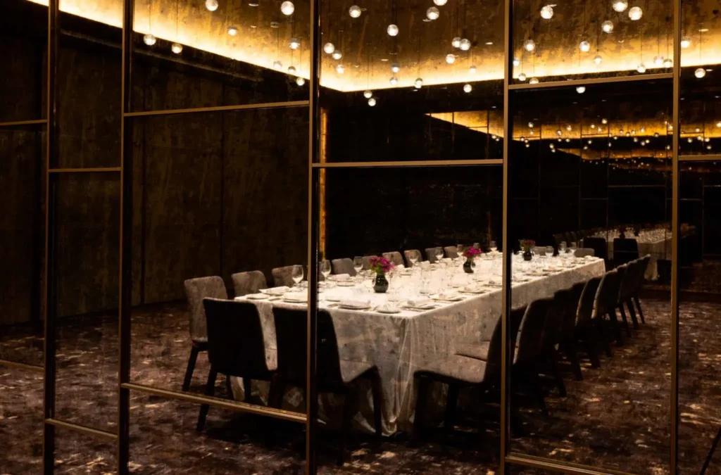 Ein langer Esstisch, der für eine formelle Veranstaltung gedeckt ist, steht in einem schwach beleuchteten Raum mit verspiegelten Wänden und Hängelampen, der sich perfekt für elegante Veranstaltungen oder intime Treffen eignet.