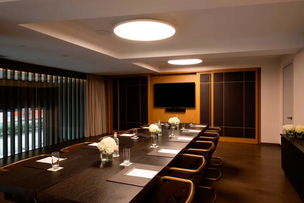 A modern conference room designed for meetings or events, featuring a long table with twelve chairs, notepads, pens, glasses of water, and white flower centerpieces, all brightly lit by ceiling lights.