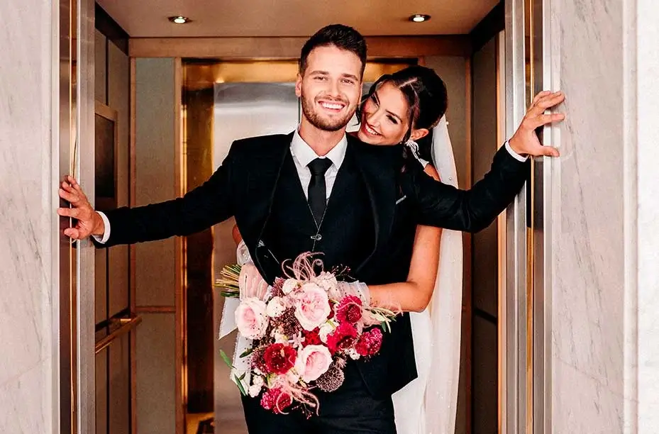 A bride and groom in formal wedding attire stand smiling inside an elevator, as if on their way to a special meeting; the groom holds a bouquet of pink and red flowers, marking the start of their unforgettable event.