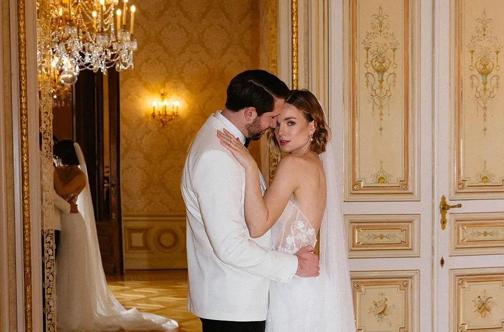 A bride and groom embrace in an elegant, ornate room with gold-trimmed doors and a chandelier during their special event, while their reflection appears in a nearby mirror.