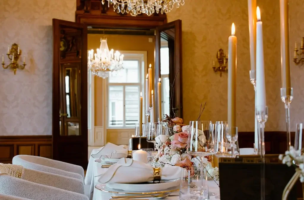 A formal dining table is set for events or a meeting, featuring white plates, glassware, floral centerpieces, and tall lit candles in an elegant, chandelier-lit room.