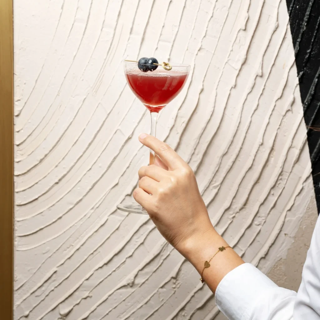 A hand holds a coupe glass filled with a red cocktail, garnished with blueberries and a lemon twist, against a textured white wall at Donnersmarkt Bar.