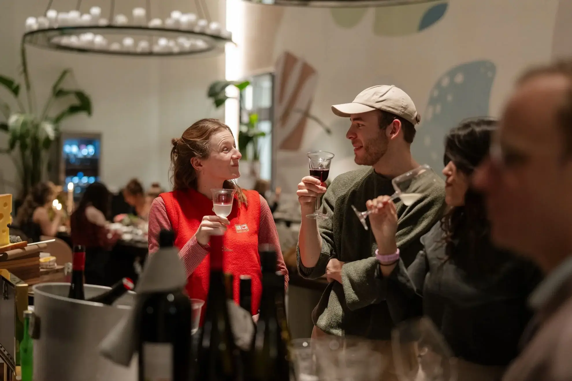 Three people stand at a bar holding glasses of wine, engaged in conversation after visiting the Thursday Markets; other patrons are visible in the background.