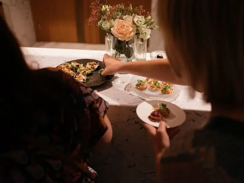 Two people select appetizers from a serving tray on a marble counter, showcasing elegant event catering with a floral arrangement in the background.