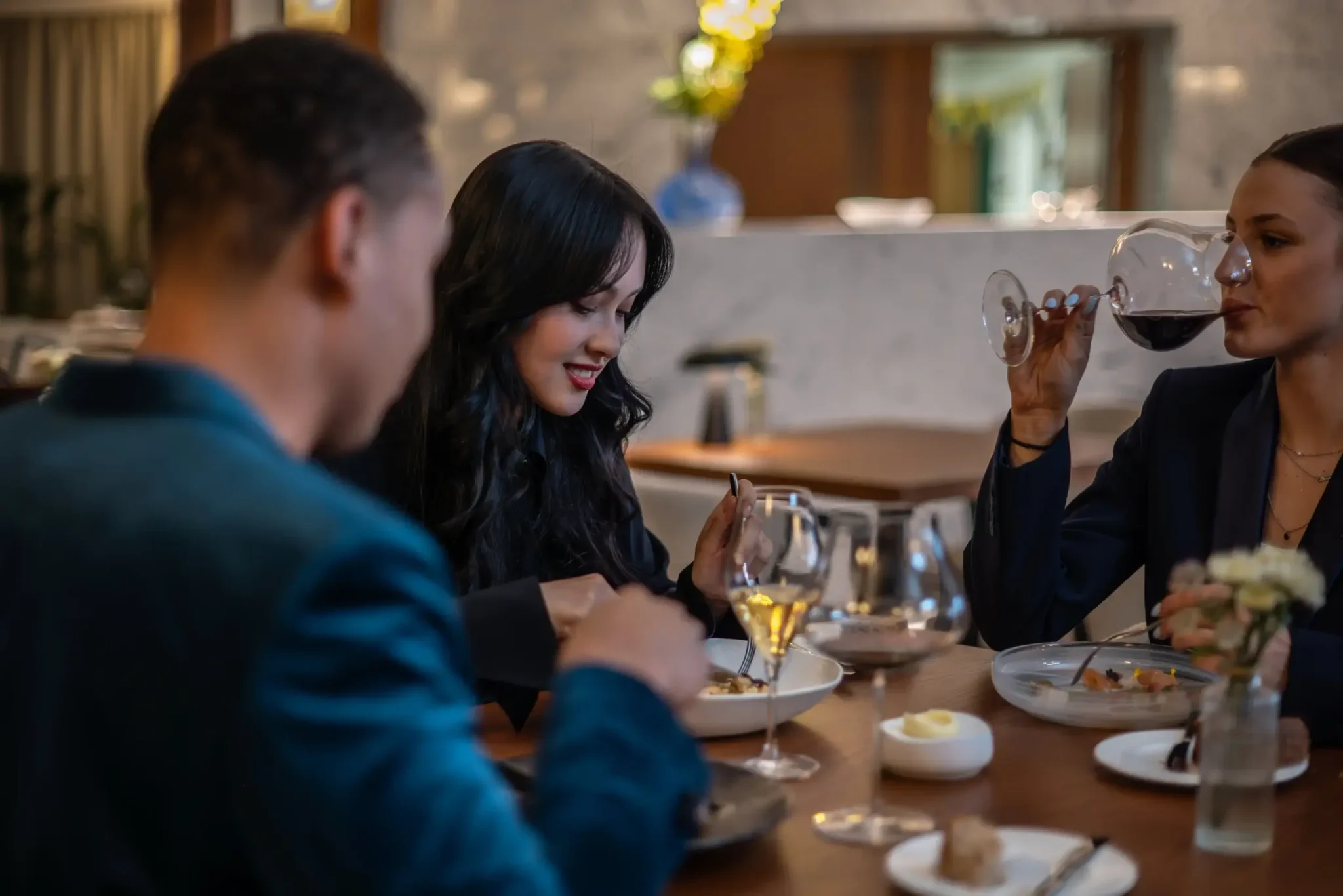 Tři lidé sedí u stolu v restauraci Restaurace Alcron, vychutnávají si víno a živě konverzují, na dřevěném stole jsou naaranžovány talíře s jídlem a skleničky.