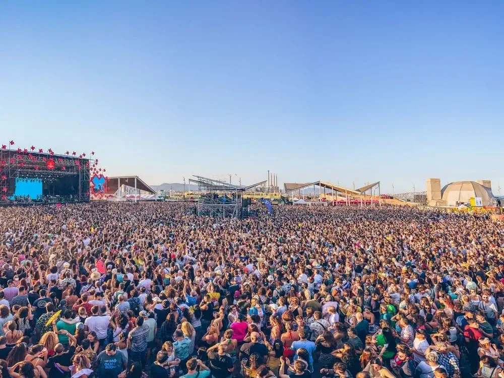 A large crowd gathered in front of an outdoor concert stage under a clear blue July sky, with event structures and distant buildings visible in the background.