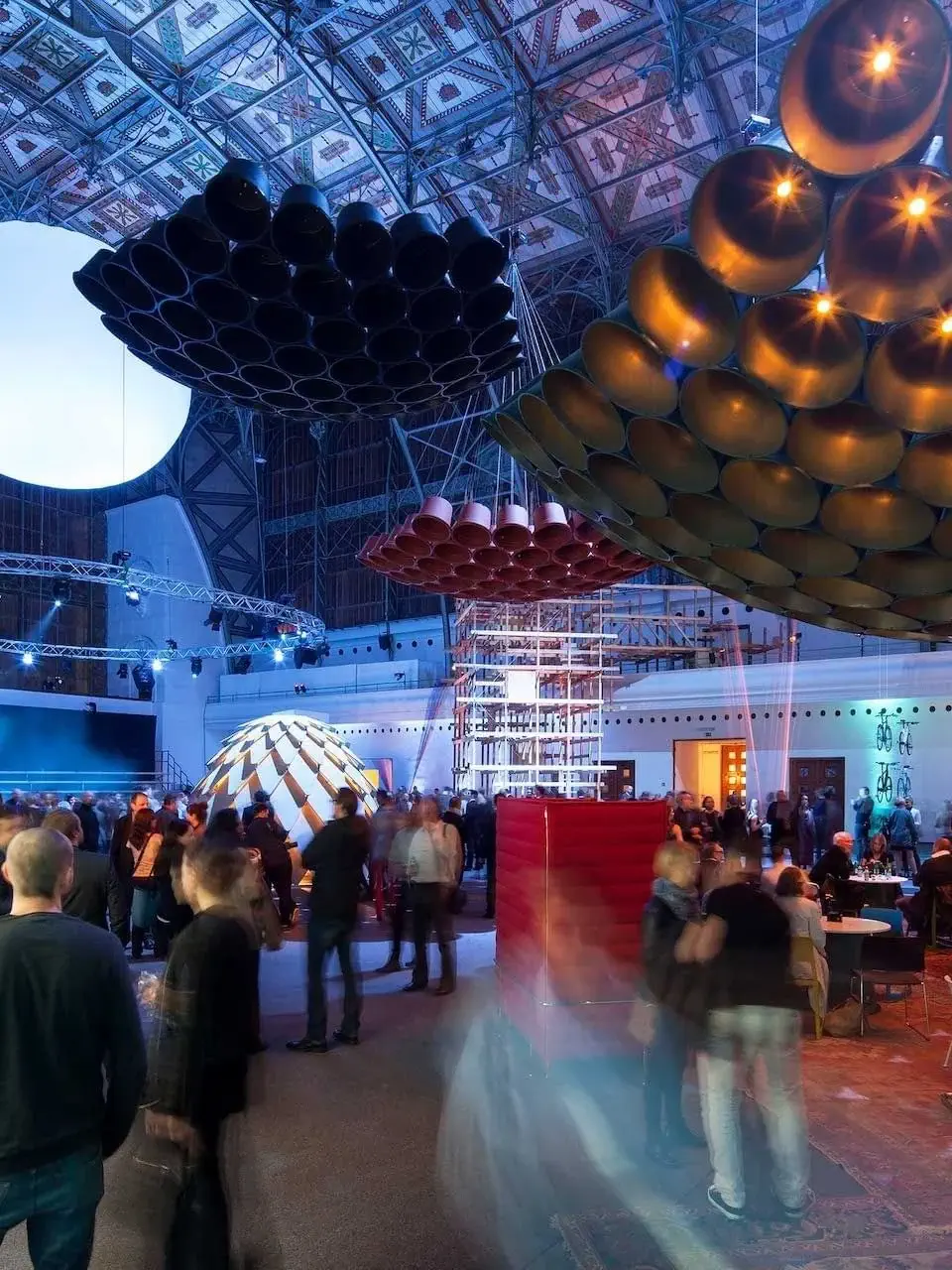 A crowd gathers in a large indoor space with geometric ceiling decor and colorful lighting at this Design Hideaway, featuring hanging sculptural installations and various inviting seating areas.