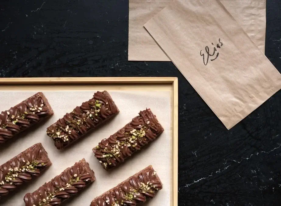 Obdélníkové čokoládové zákusky s polevou a ořechovou polevou jsou na tmavém podnose naaranžovány vedle hnědých papírových sáčků s nápisem "Elis".