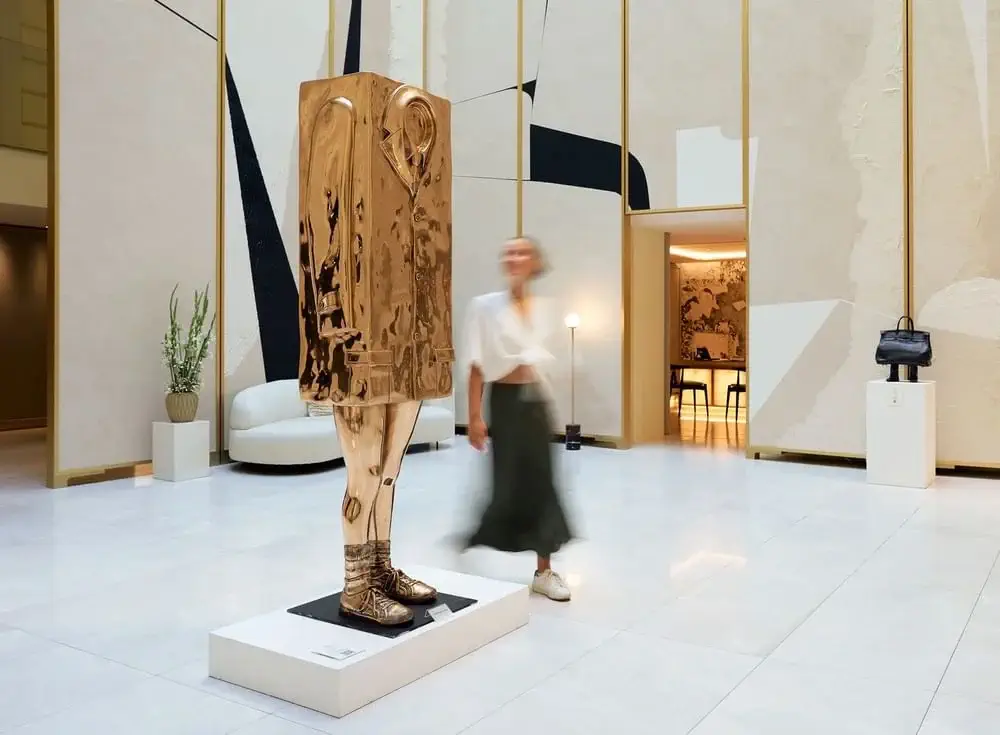 Eine Frau geht an einer modernen, abstrakten Skulptur einer Figur mit einem rechteckigen goldenen Kopf und Torso vorbei, die in einer hellen, minimalistischen Lobby ausgestellt ist.