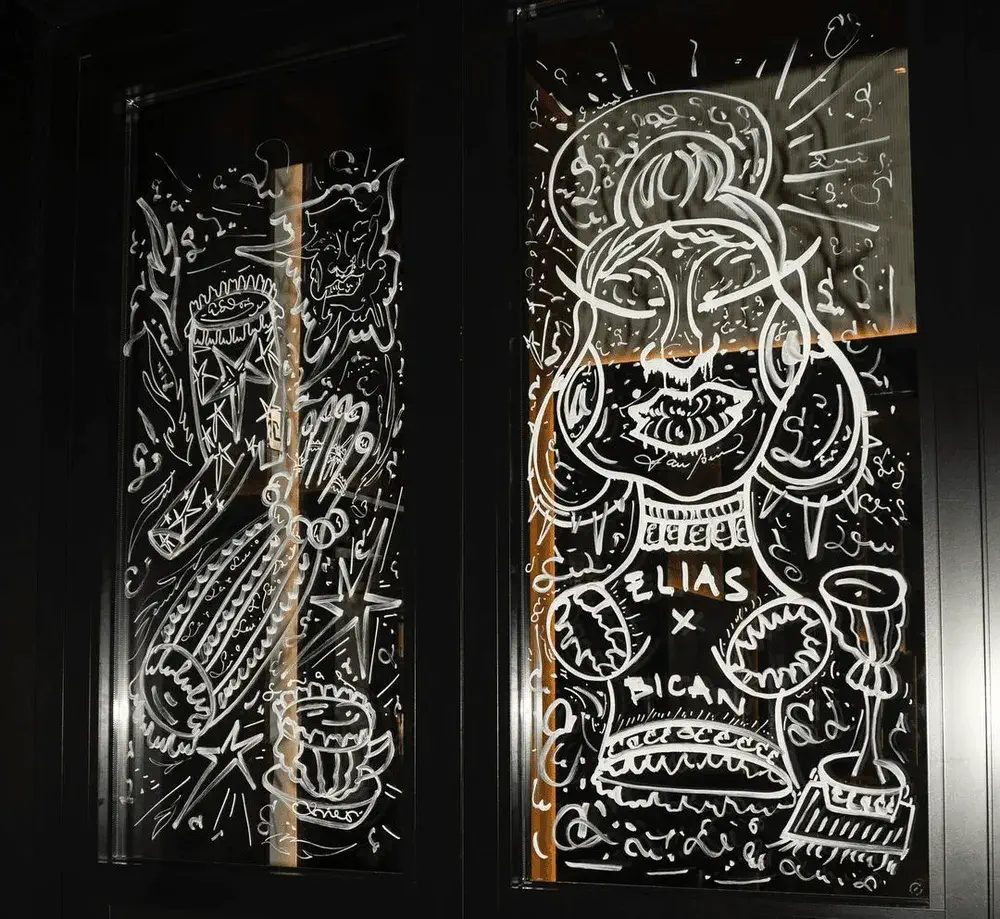 White line art drawings on glass panels depict a stylized figure with "ELIAS PICAN" written on the chest, surrounded by musical instruments and abstract designs.