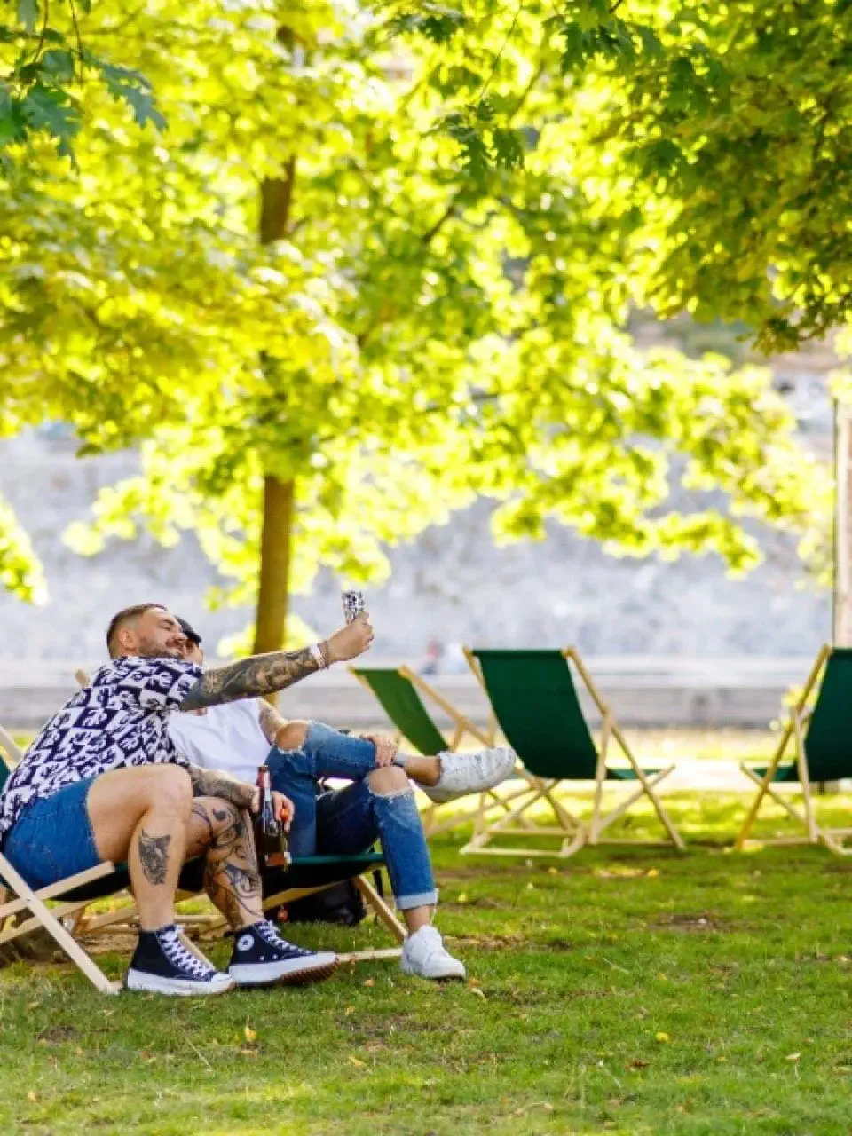 Two people sit on deck chairs in a Prague park, taking a selfie under leafy trees on a sunny day, with more empty chairs in the background.