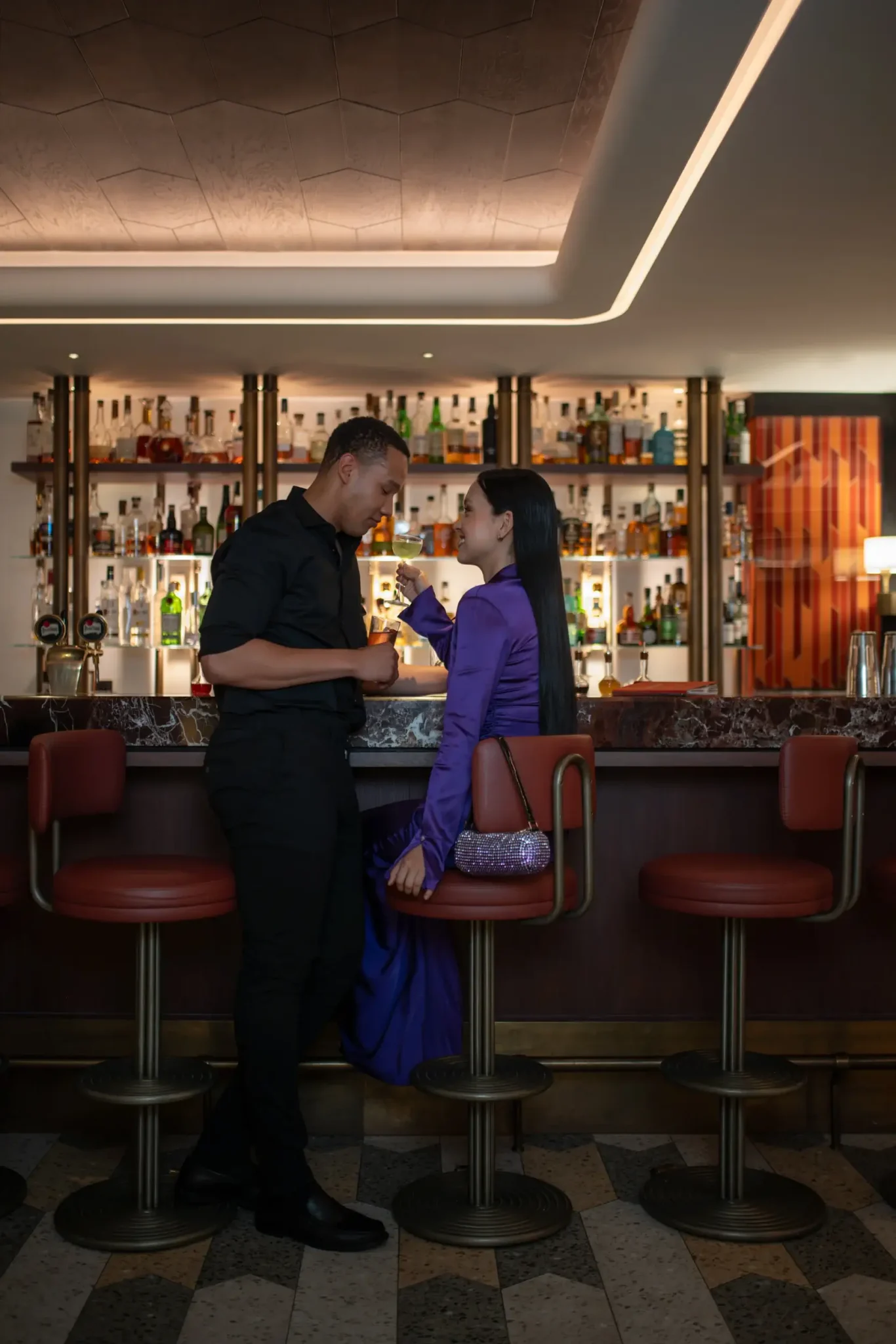 Ein Mann und eine Frau unterhalten sich an einer Bar; die Frau sitzt auf einem Hocker in einem lila Kleid, während der Mann ihr gegenübersteht. An der Rückwand stehen Regale mit Flaschen.