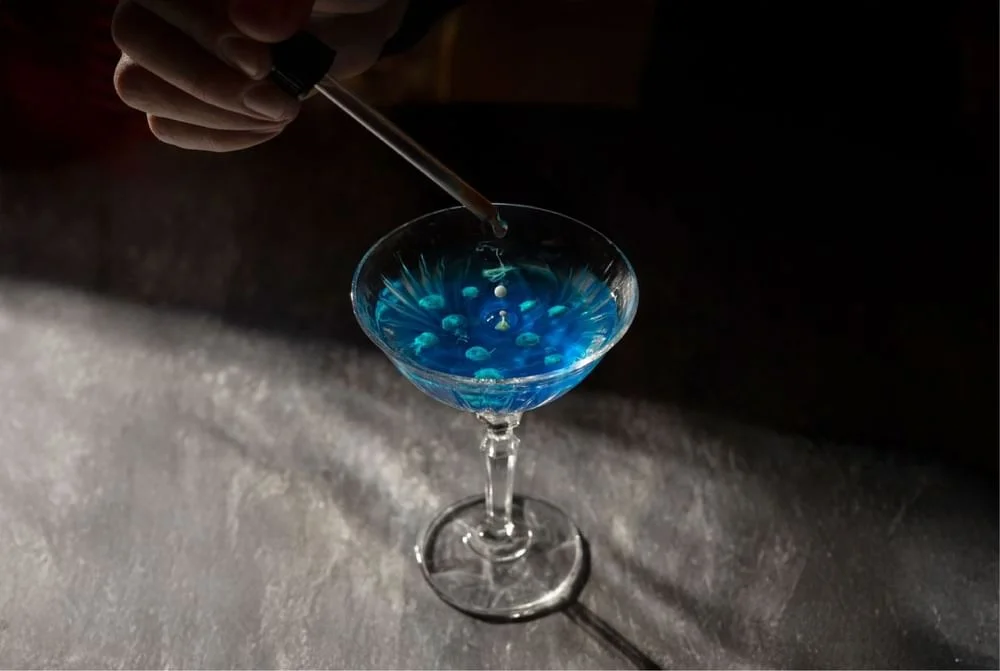 Una mà utilitza un comptagotes per afegir líquid en un got de còctel Terrassa Azimuth ple de líquid blau sobre una superfície fosca.