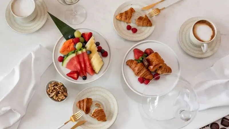 A breakfast table set with coffee cups, croissants, a plate of assorted fresh fruit, walnuts, and fresh berries on white dishes with gold forks.