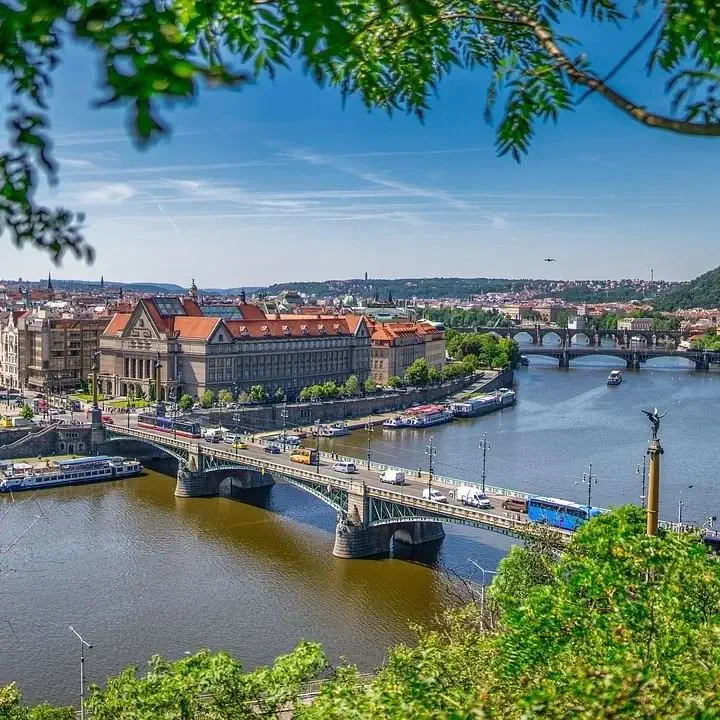 Letecký pohled na město PRAHA v KVĚTENĚ, s řekou vinoucí se kolem několika mostů, historickými budovami s červenými střechami a bujnou zelení v popředí pod jasně modrou oblohou.